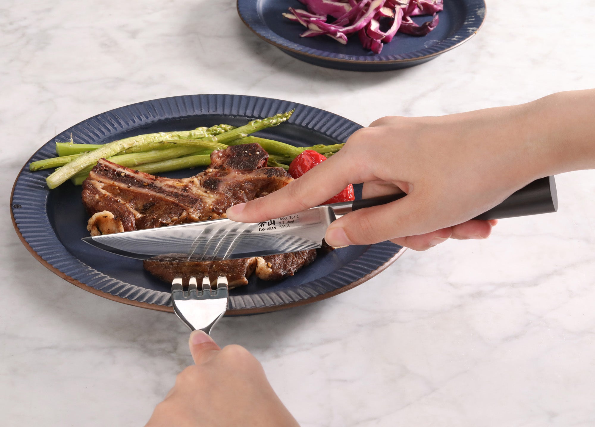 Hand holding a steak knife cutting a cooked steak on a plate with asparagus and a fork.
