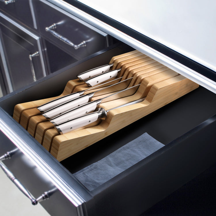 Seven stainless steel knives with white handles arranged in a bamboo in-drawer knife organizer inside a kitchen drawer.