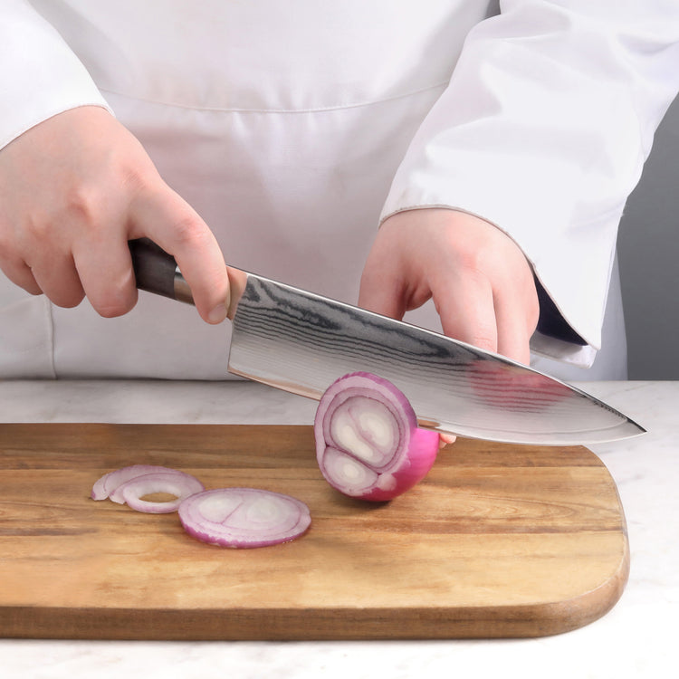 Hands holding a Damascus steel knife slicing a red onion on a wooden cutting board.