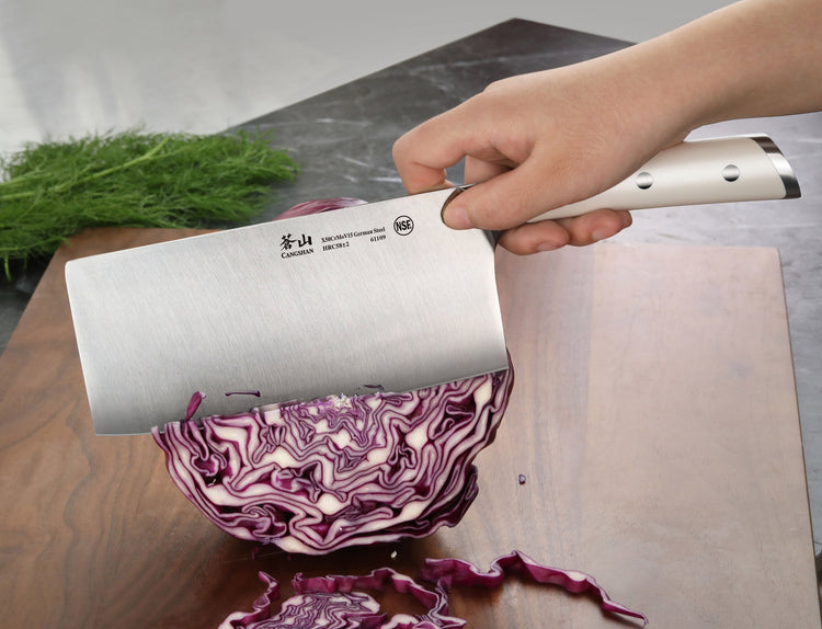 Hand holding a cleaver knife cutting a halved purple cabbage on a wooden cutting board with herbs in the background.