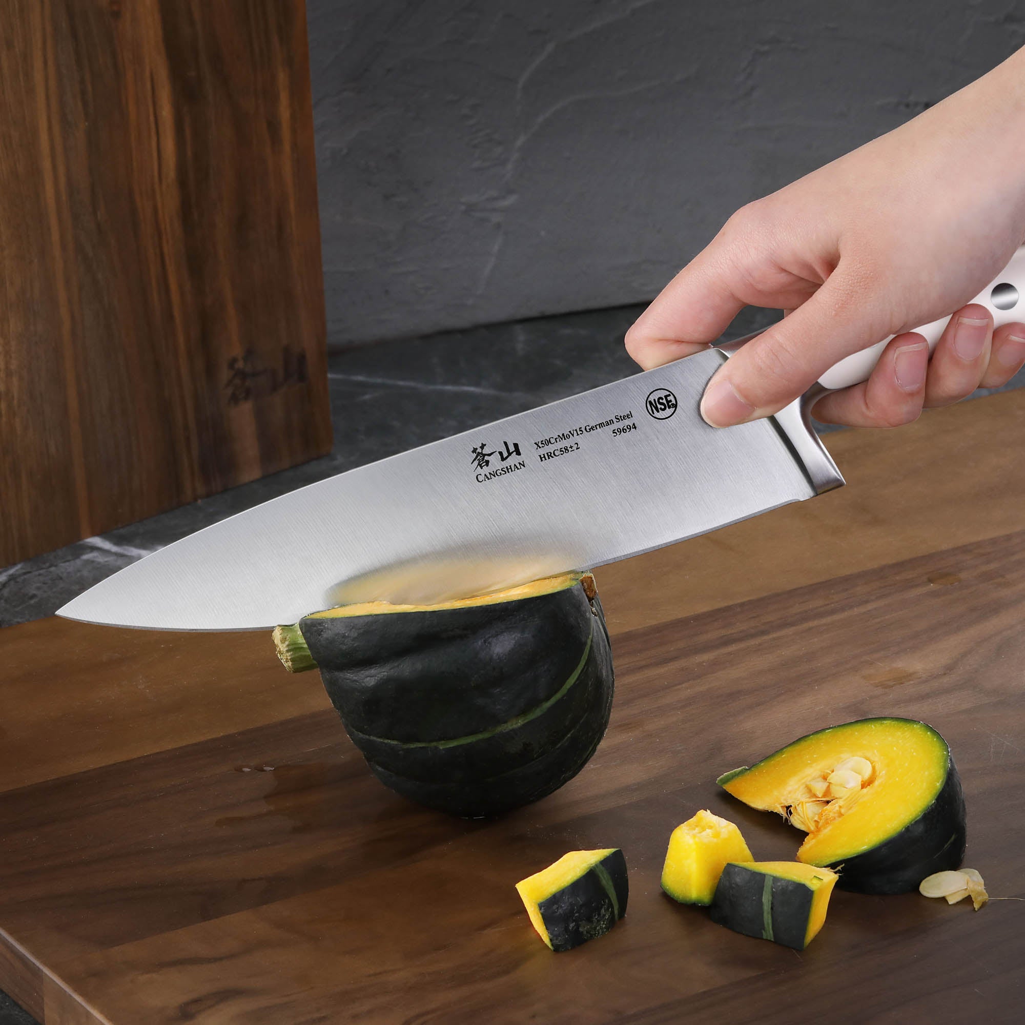 Hand holding a large cleaver knife cutting a small green and yellow squash on a wooden cutting board with a wooden knife bloc