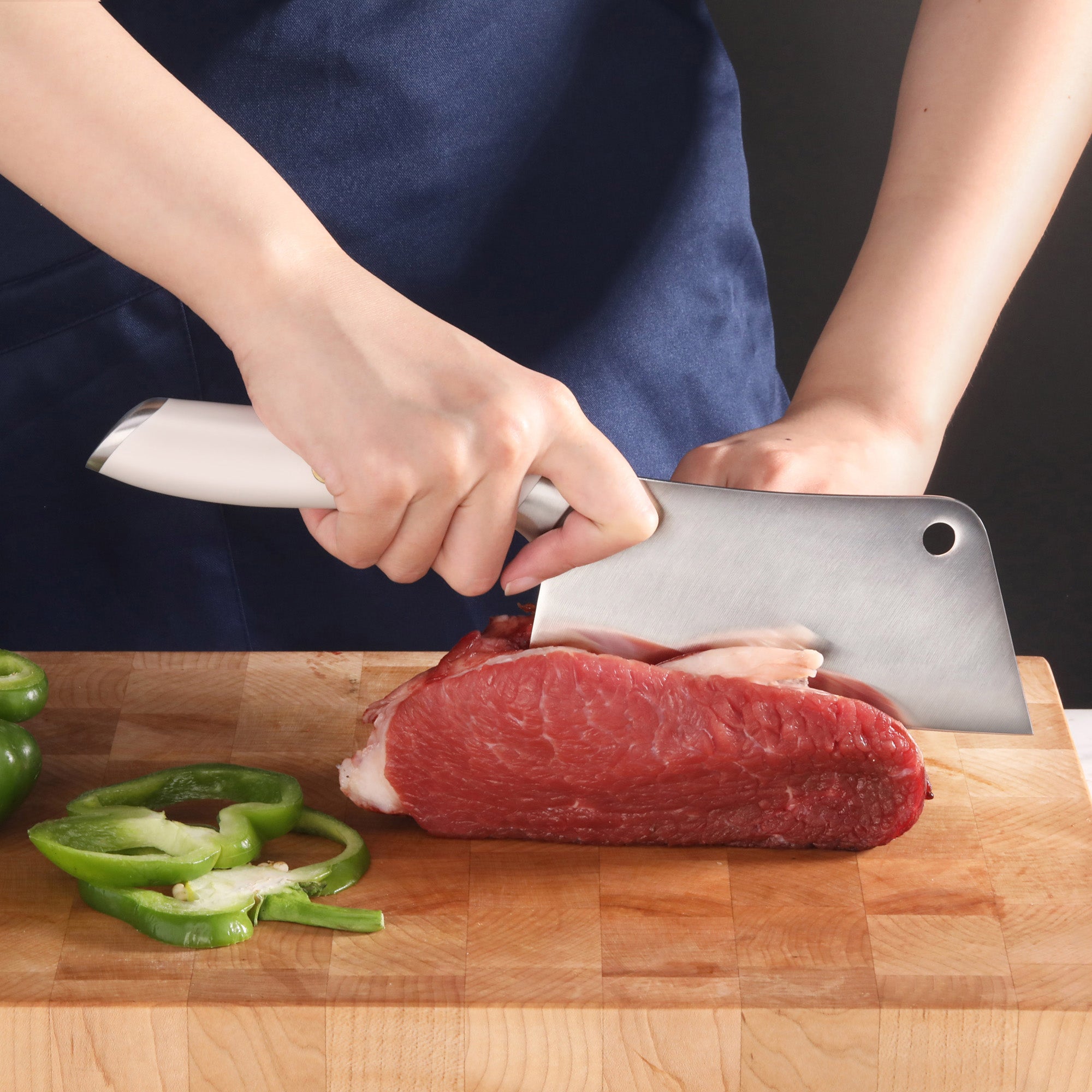 Hand holding a cleaver knife slicing raw beef on a wooden cutting board with sliced green bell peppers nearby.
