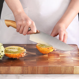 
                    
                      Hand holding a knife with a wooden handle, slicing a horned melon on a wooden cutting board.
                    
                  
