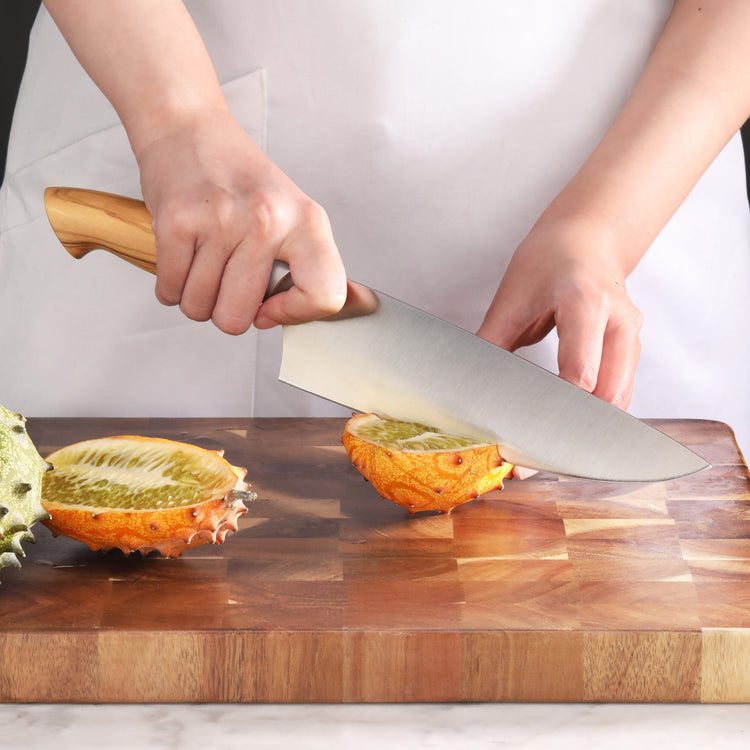 Hand holding a knife with a wooden handle, slicing a horned melon on a wooden cutting board.