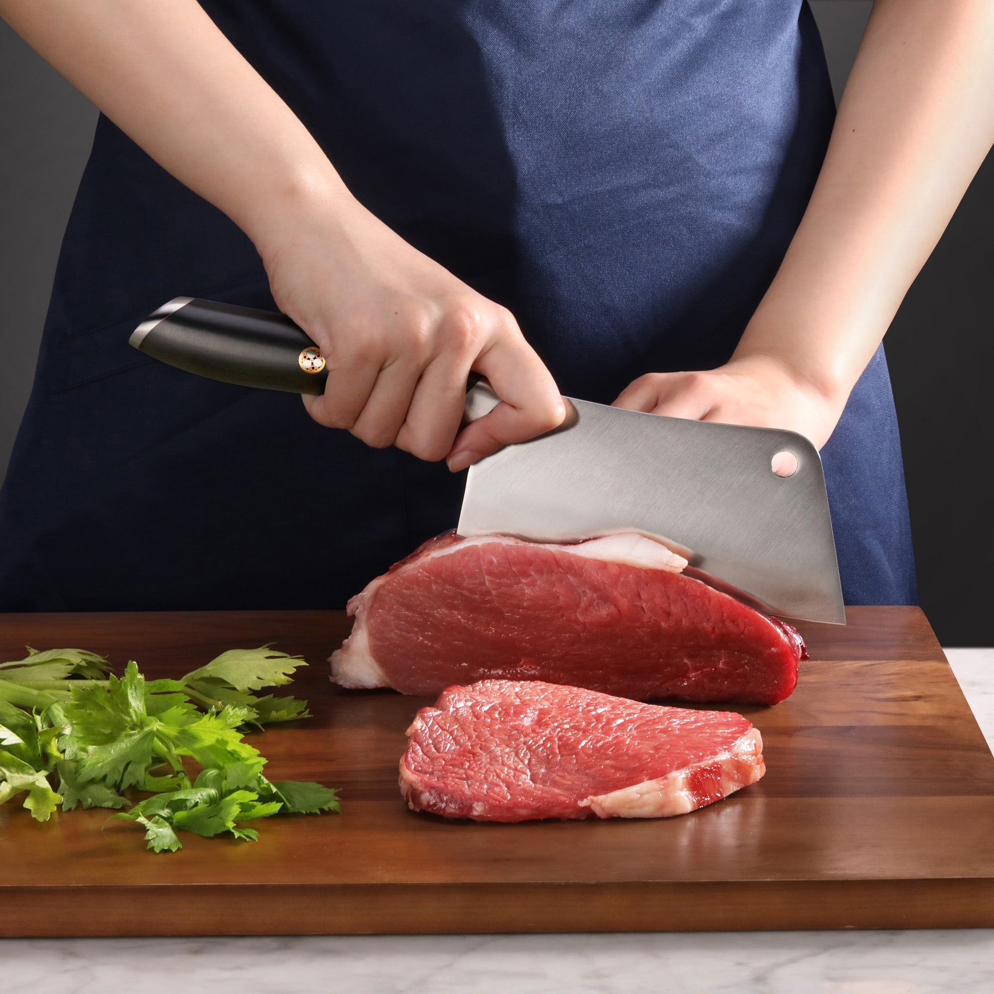 Person slicing raw beef on wooden cutting board with large cleaver knife, fresh herbs nearby.