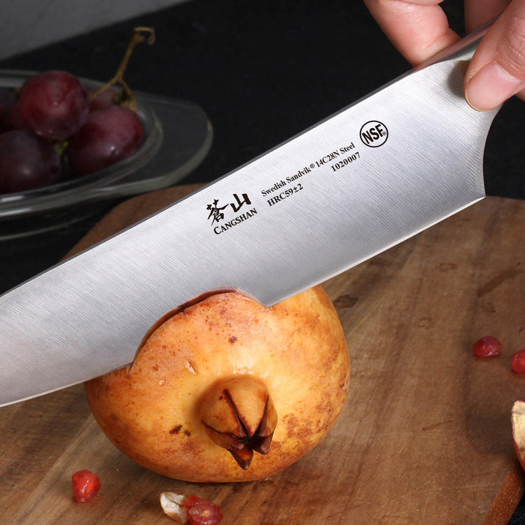 Knife blade labeled Cangshan slicing a pomegranate on a wooden cutting board with grapes in the background.