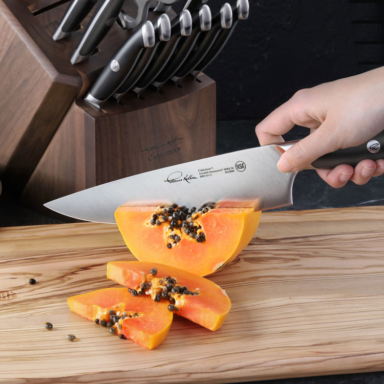 Hand holding a chef's knife slicing a papaya on a wooden cutting board with a knife block in the background.