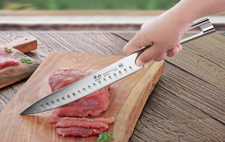 Hand holding a carving knife slicing raw meat on a wooden cutting board outdoors.
