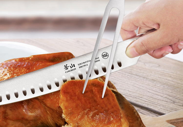 Hand holding fork lifting slice of roasted meat while knife with holes slices it on wooden surface.