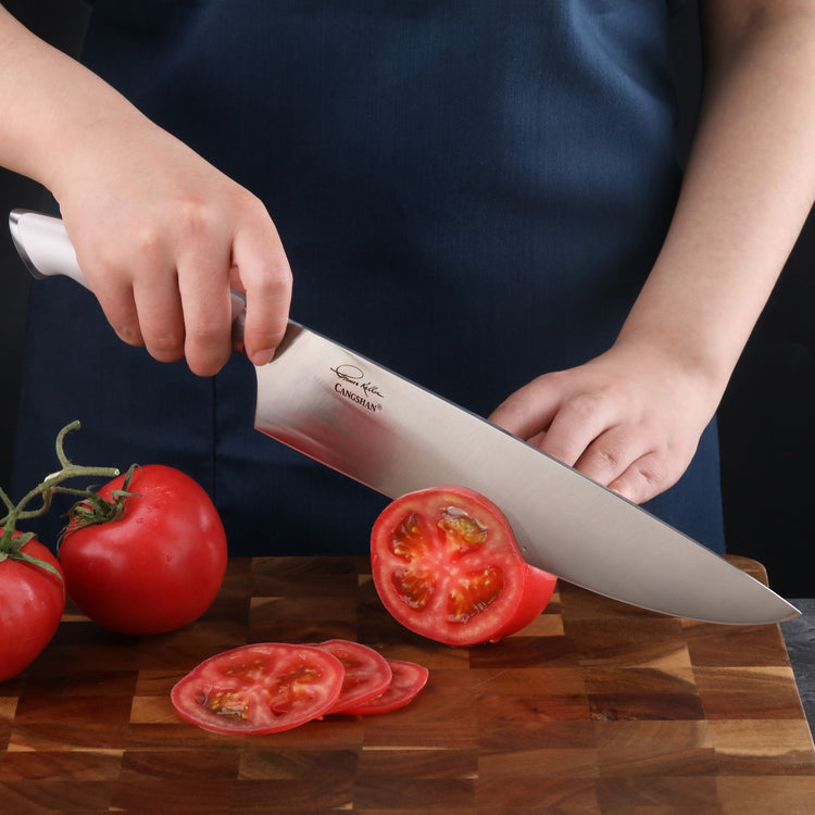 Person slicing a tomato on a wooden cutting board with a large stainless steel chef's knife.