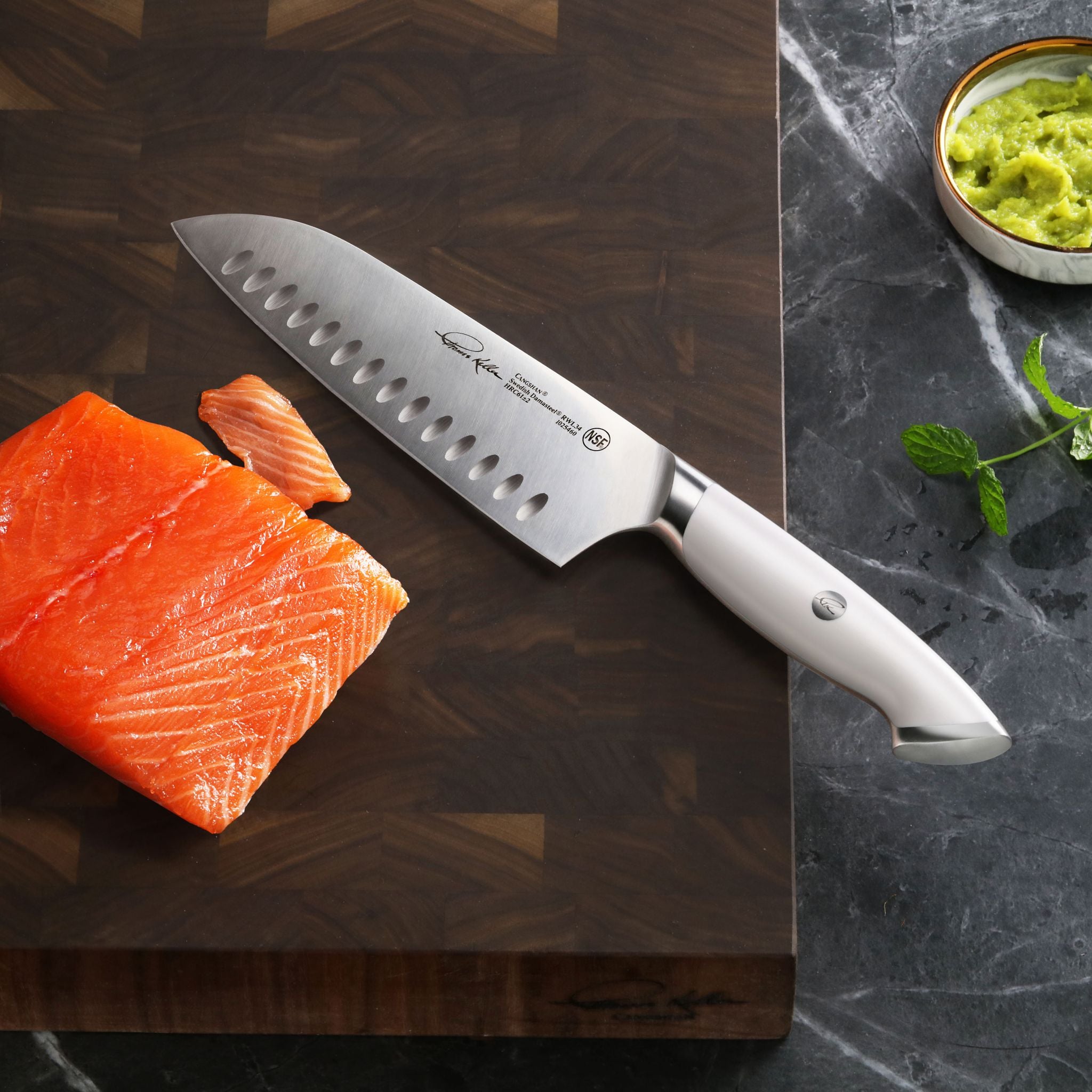 TKSC 7-inch Santoku knife with white handle on wooden cutting board next to sliced salmon and bowl of green sauce.