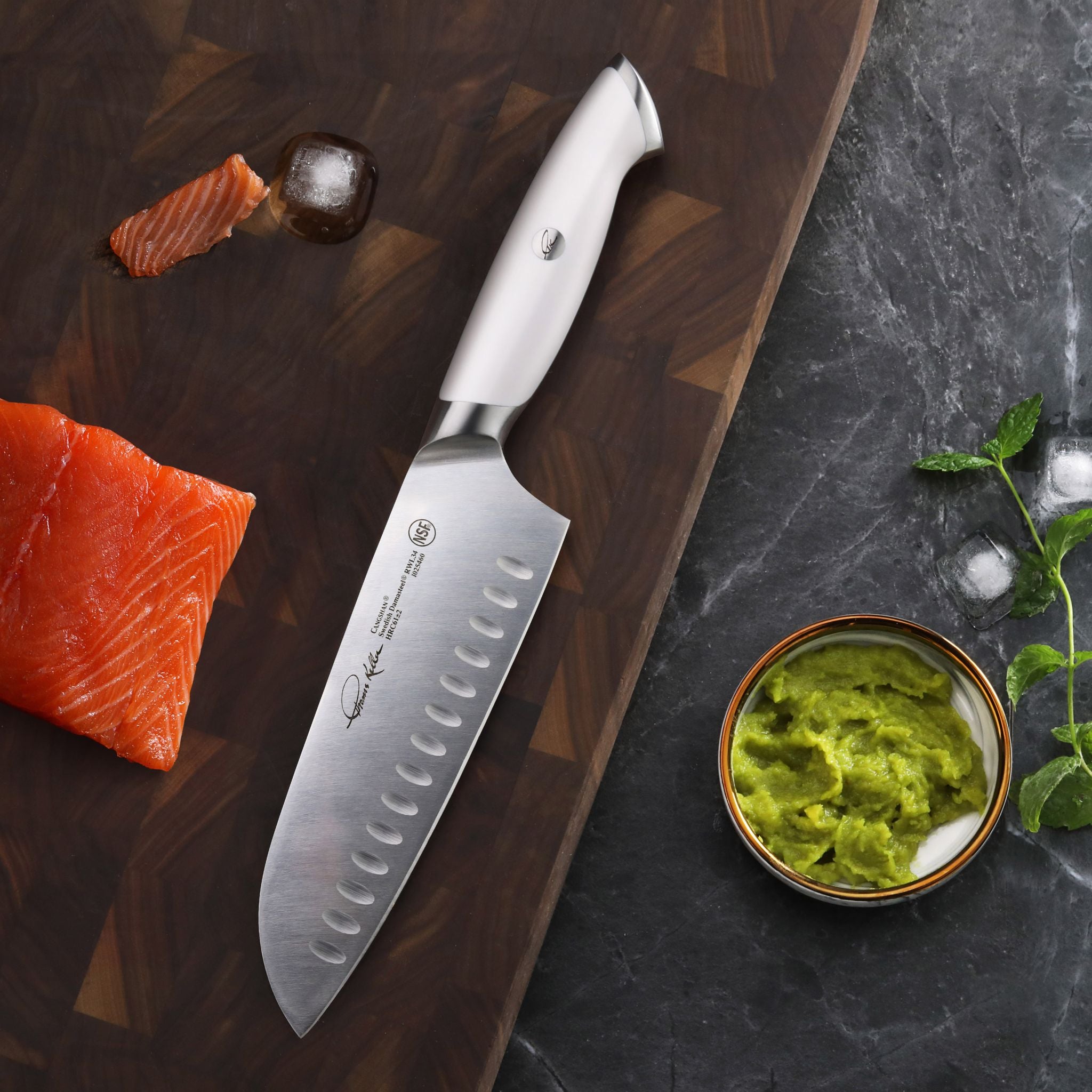 7-inch Santoku knife with white handle on wooden cutting board next to salmon and bowl of green sauce.