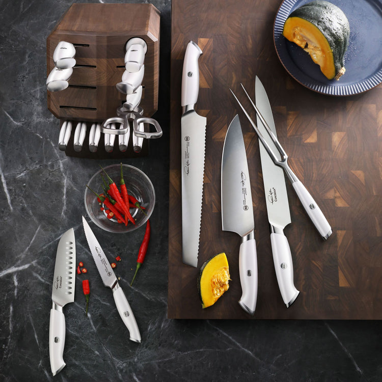 Set of white-handled kitchen knives, honing steel, and fork on wooden cutting board with sliced squash and chili peppers near