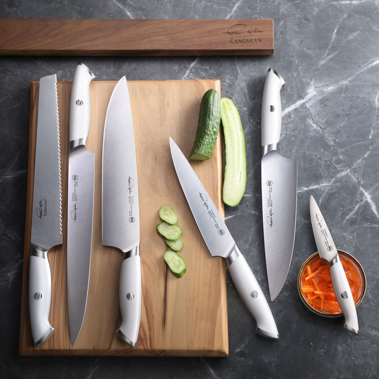 Seven white-handled knives with various blade shapes arranged on and beside a wooden cutting board with sliced cucumber and a