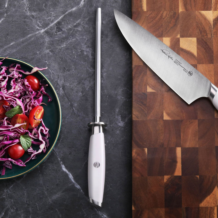 White-handled honing steel with stainless steel rod, chef knife on wooden cutting board, and bowl of salad on dark countertop