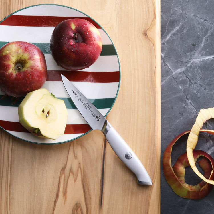 Paring knife with white handle on a striped plate with two whole apples and one sliced apple, next to apple peels.