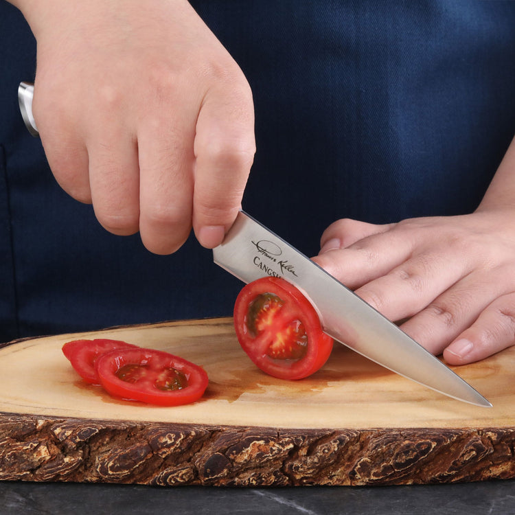 Hand holding a utility knife slicing a tomato on a wooden cutting board.