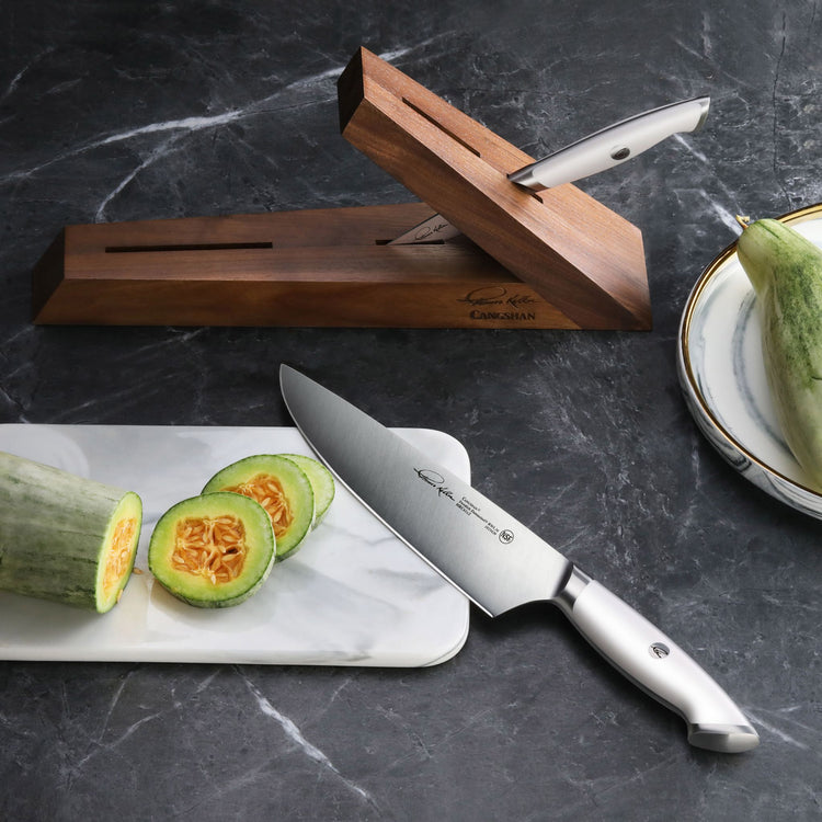 Walnut knife block with two knives, one inserted and one on marble cutting board slicing a green squash.