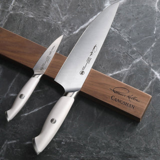 
                    
                      Two stainless steel knives with white handles resting on a walnut magnetic knife bar on a gray marble surface.
                    
                  