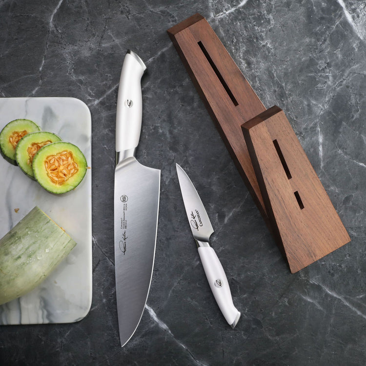 Two knives with white handles and a wooden knife block on a dark marble surface next to sliced cucumber on a marble cutting b