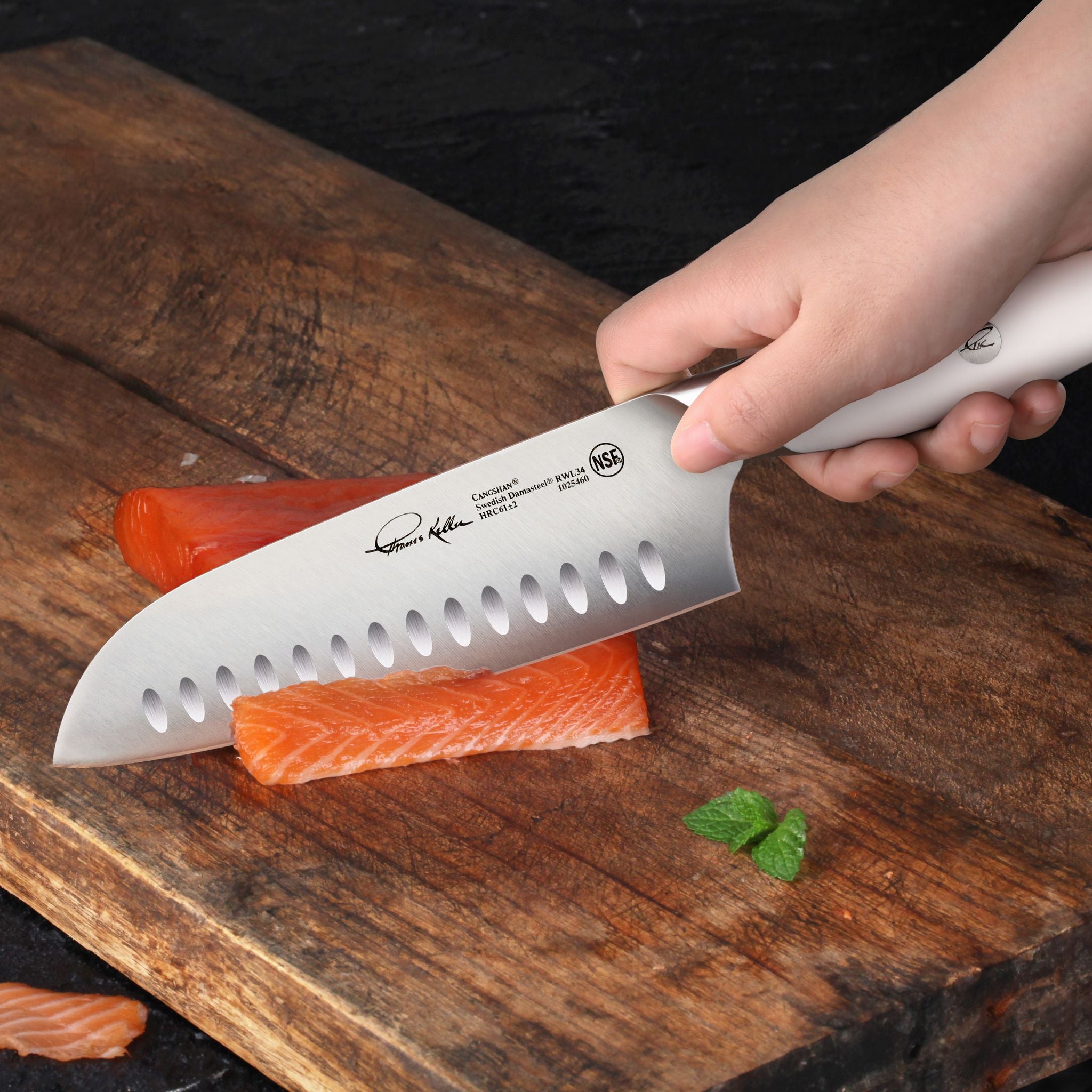 Hand holding a white-handled Santoku knife slicing raw salmon on a wooden cutting board with a small green garnish nearby.